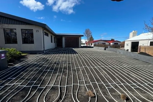 Bílaplan við íbúðarhús þakið hvítum snjóbræðslulögnum sem lagðar eru í þéttum lykkjum á malarundirlag, heiðskír himinn.