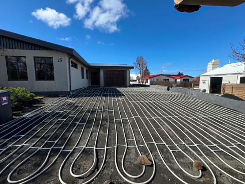 Bílaplan við íbúðarhús þakið hvítum snjóbræðslulögnum sem lagðar eru í þéttum lykkjum á malarundirlag, heiðskír himinn.