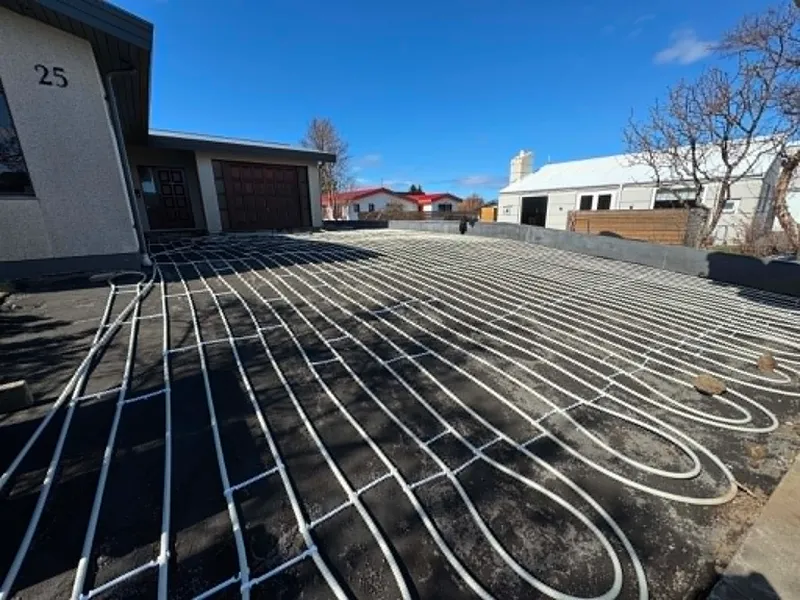 Bílaplan við hús númer 25 með snjóbræðslulögnum í lykkjum á malarundirlagi, bílskúrshurð og nágrannahús í bakgrunni.