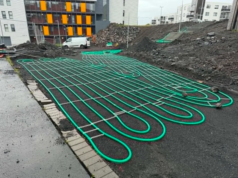 Bílastæði við Helgafellsskóla þakið grænum snjóbræðslulögnum í þéttum lykkjum á svörtu undirlagi, íbúðarhús og sendibíll í bakgrunni.