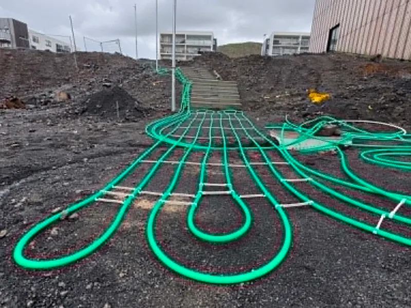 Snjóbræðslulagnir liggja í lykkjum upp að steyptum tröppum í brekku, skólabygging og íbúðarhús efst í mynd.