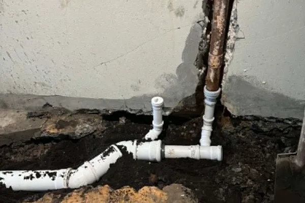 Nýjar hvítar plastlagnir með greinitengjum tengdar við gamla ryðgaða steypujárnslögn við vegg í uppgrafinni rennu.