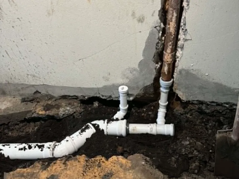 Nýjar hvítar plastlagnir með greinitengjum tengdar við gamla ryðgaða steypujárnslögn við vegg í uppgrafinni rennu.