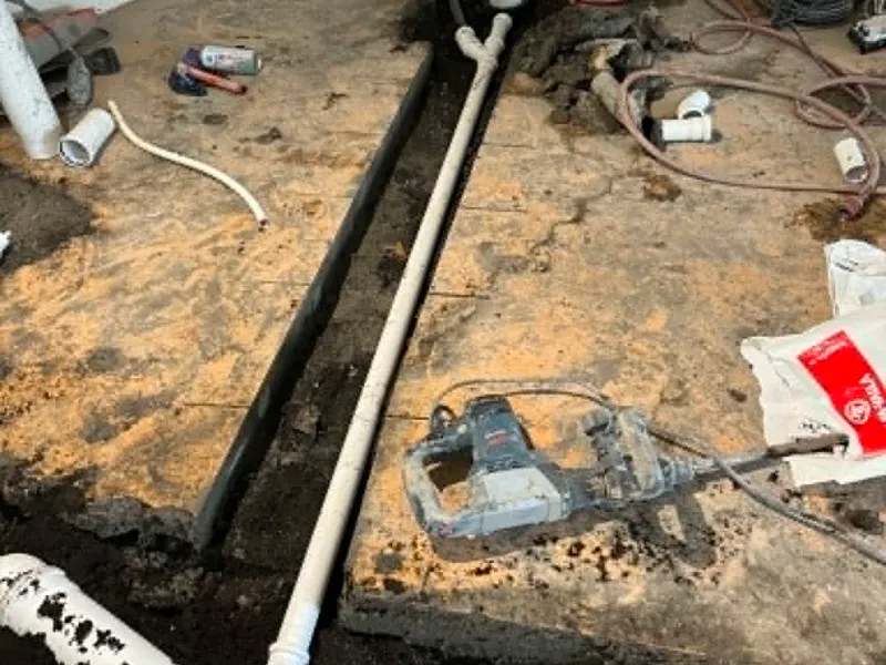 Gömul svört steypujárnslögn liggur uppbrotin á gólfi við hlið nýrrar hvítrar plastlagnar, með brotvél og gömlum tengjum.